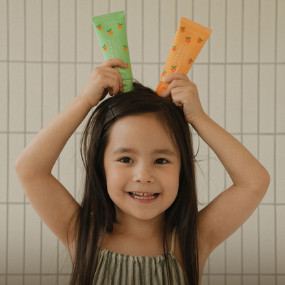 Joyful young girl holding two Unpa Cha Cha Kids Carrot Toothpaste tubes – a fun and safe toothpaste choice for kids.